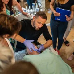 Mężczyzna w rękawiczkach wykonuje masaż kobido na twarzy kobiety. Wokół zgromadzone osoby obserwują.