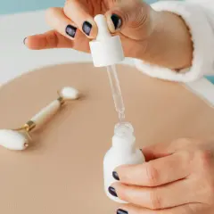 Dłoń z ciemnym lakierem do paznokci trzyma pipetkę z serum. Czy niacynamid można łączyć z kwasem hialuronowym? Obok leży jadeitowy roller.